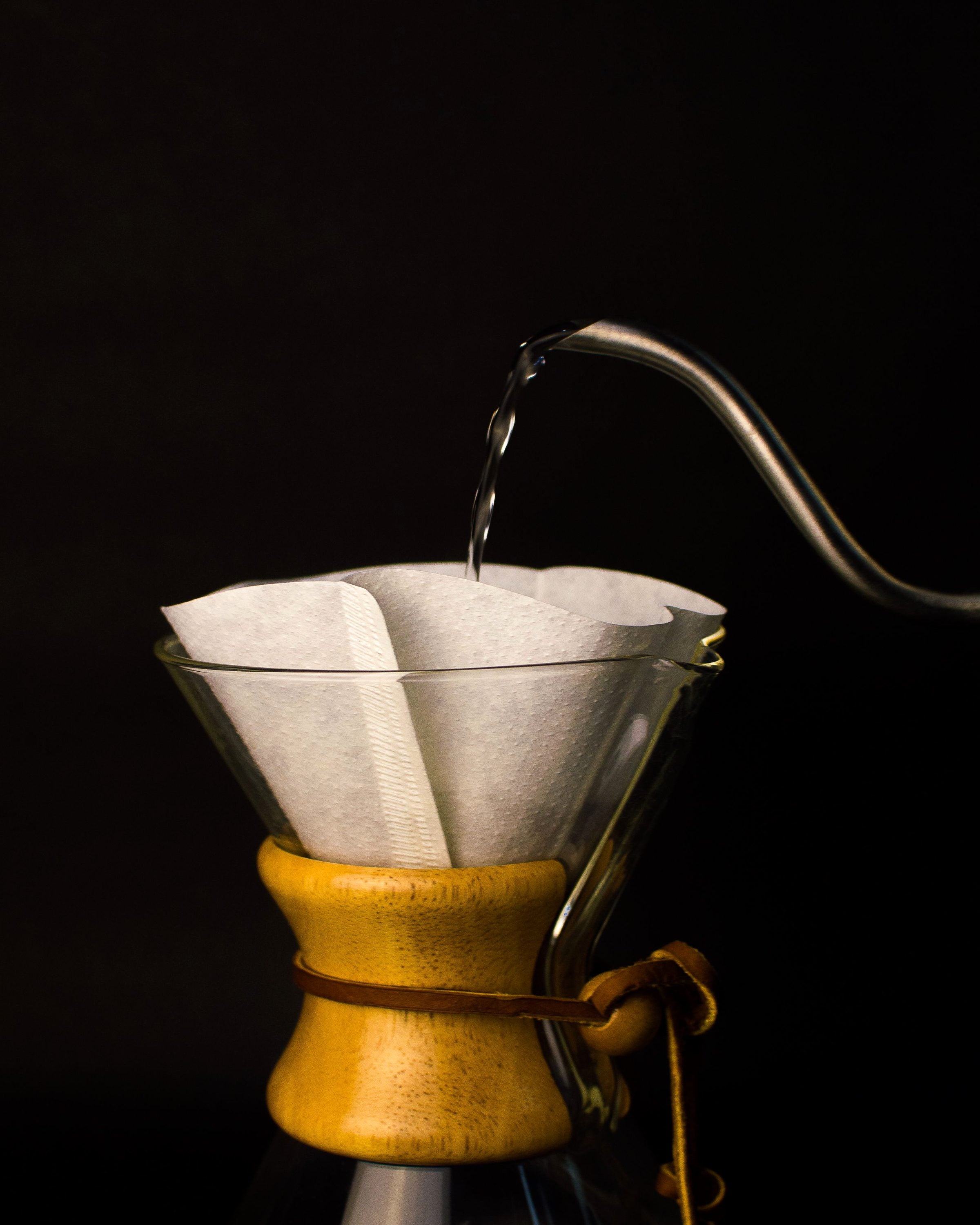 A gooseneck kettle pouring hot water over a paper filter in a Chemex, dark moody studio light
